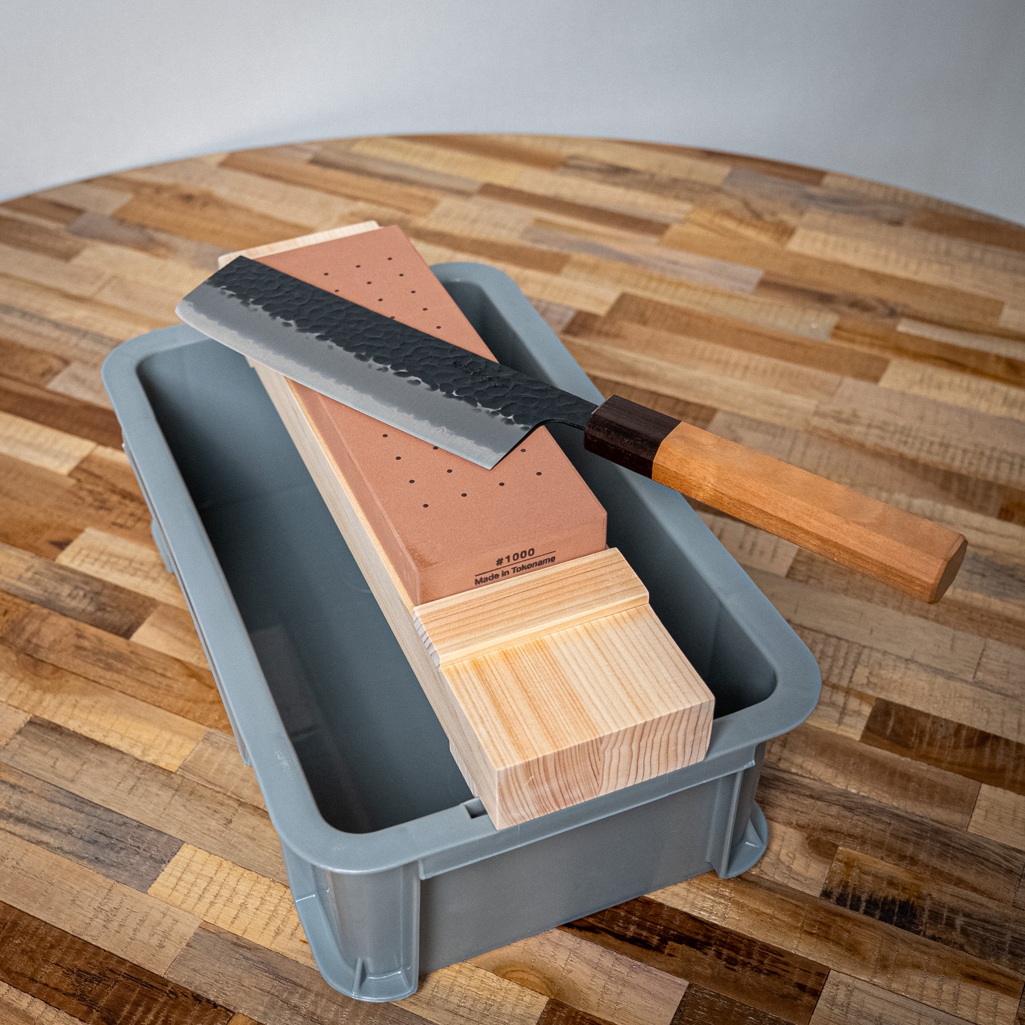 Couteau japonais sur pierre à aiguiser #1000 avec support en bois et bac gris.


