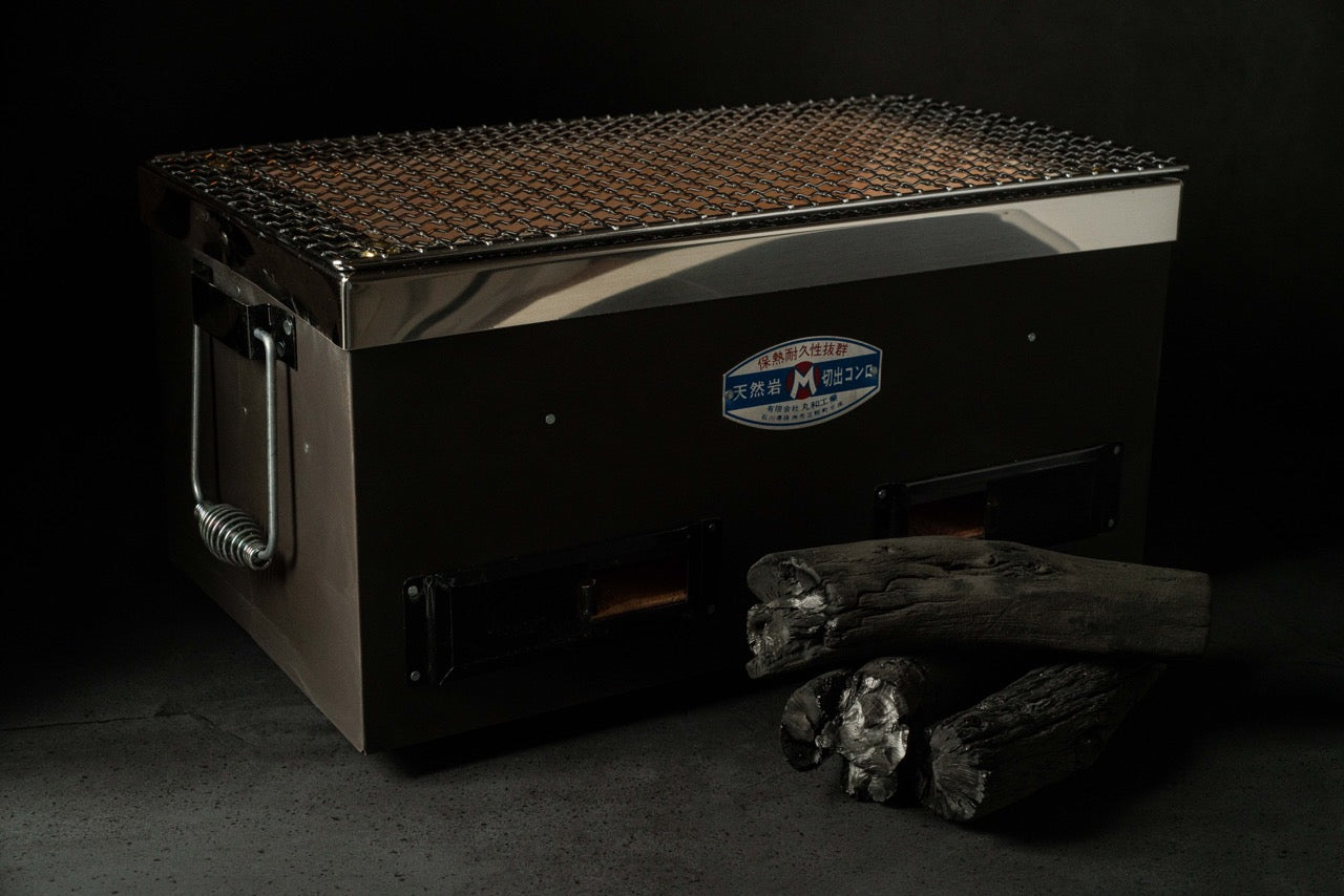 Japanese rectangular charcoal grill with metal grate and side handles, shown with pieces of charcoal on a dark background.
