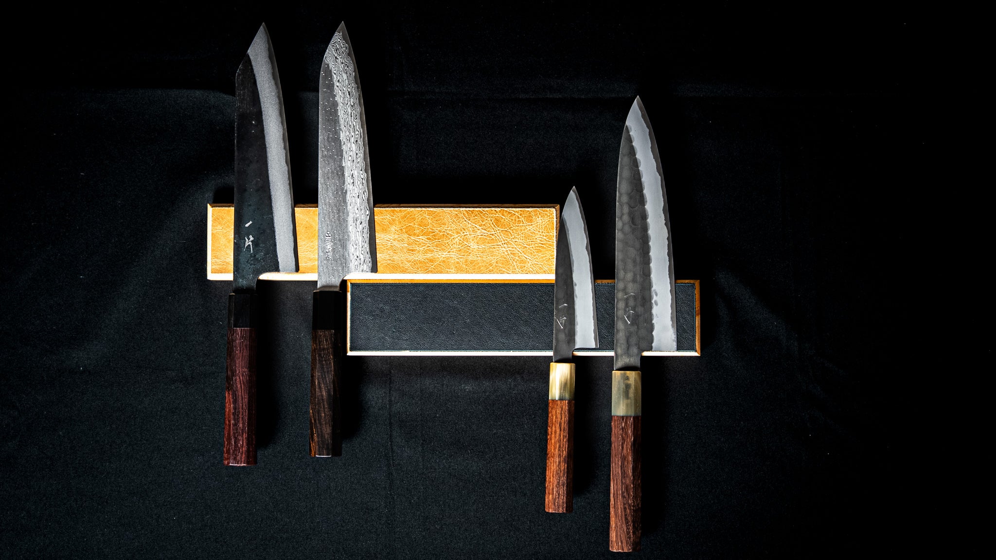 Barre magnétique murale artisanale Stay Sharp en bois et cuir noir, fixée sur un fond noir, avec deux couteaux japonais à lame martelée.