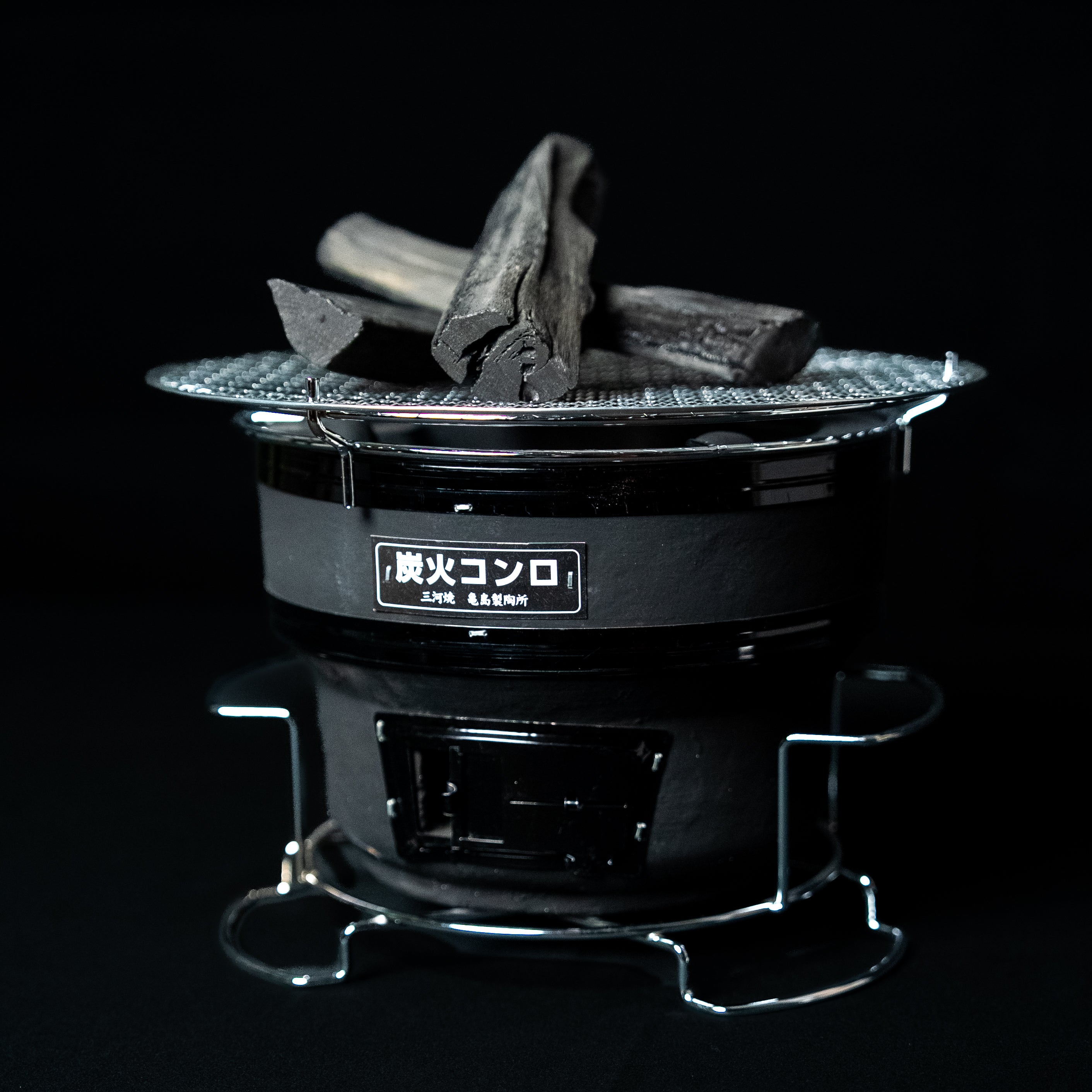 Small round Japanese charcoal grill with a metal grate and black body, shown with charcoal pieces on top against a dark background.