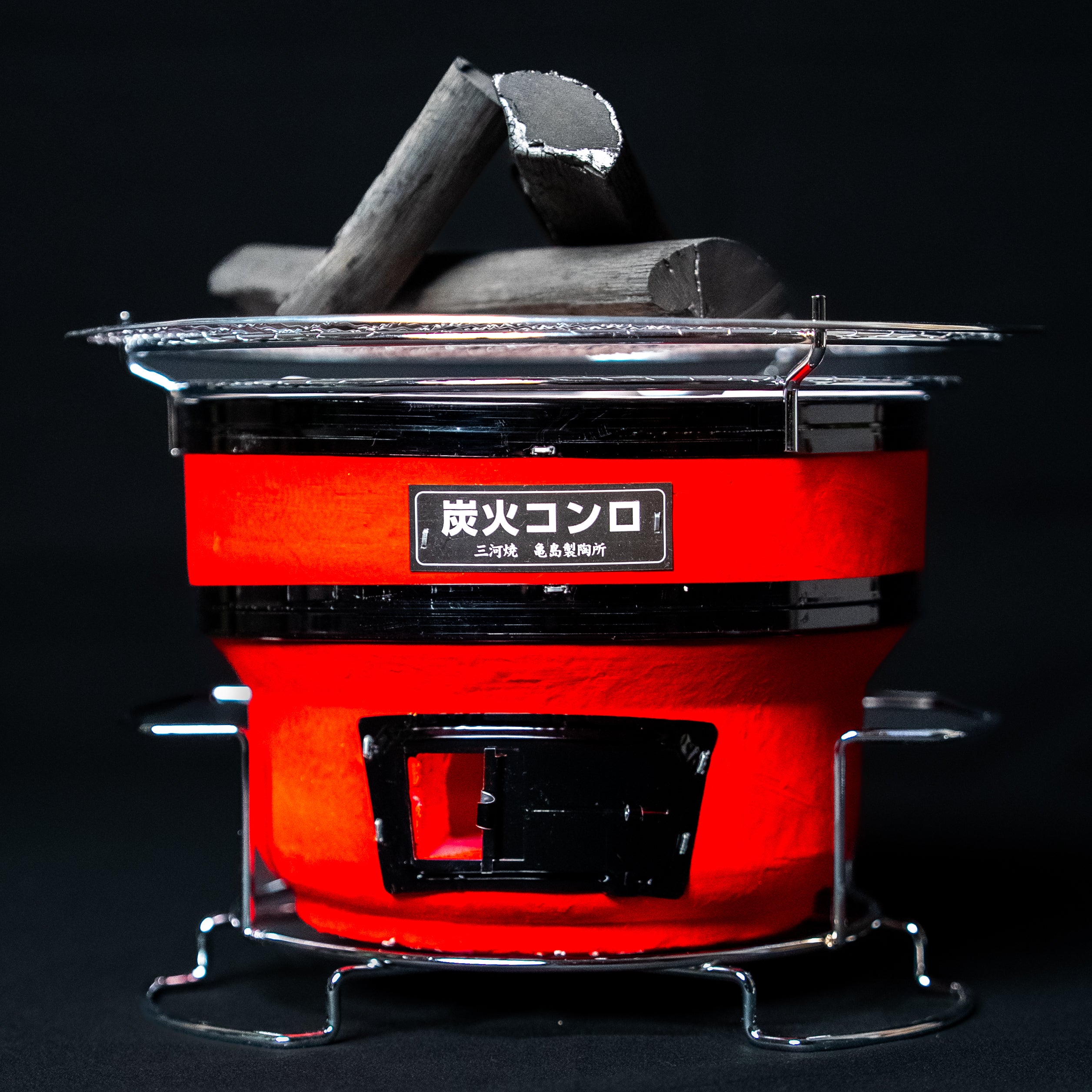 Small round Japanese charcoal grill in red with a black label and vent, topped with metal grate and charcoal pieces, against a dark background.