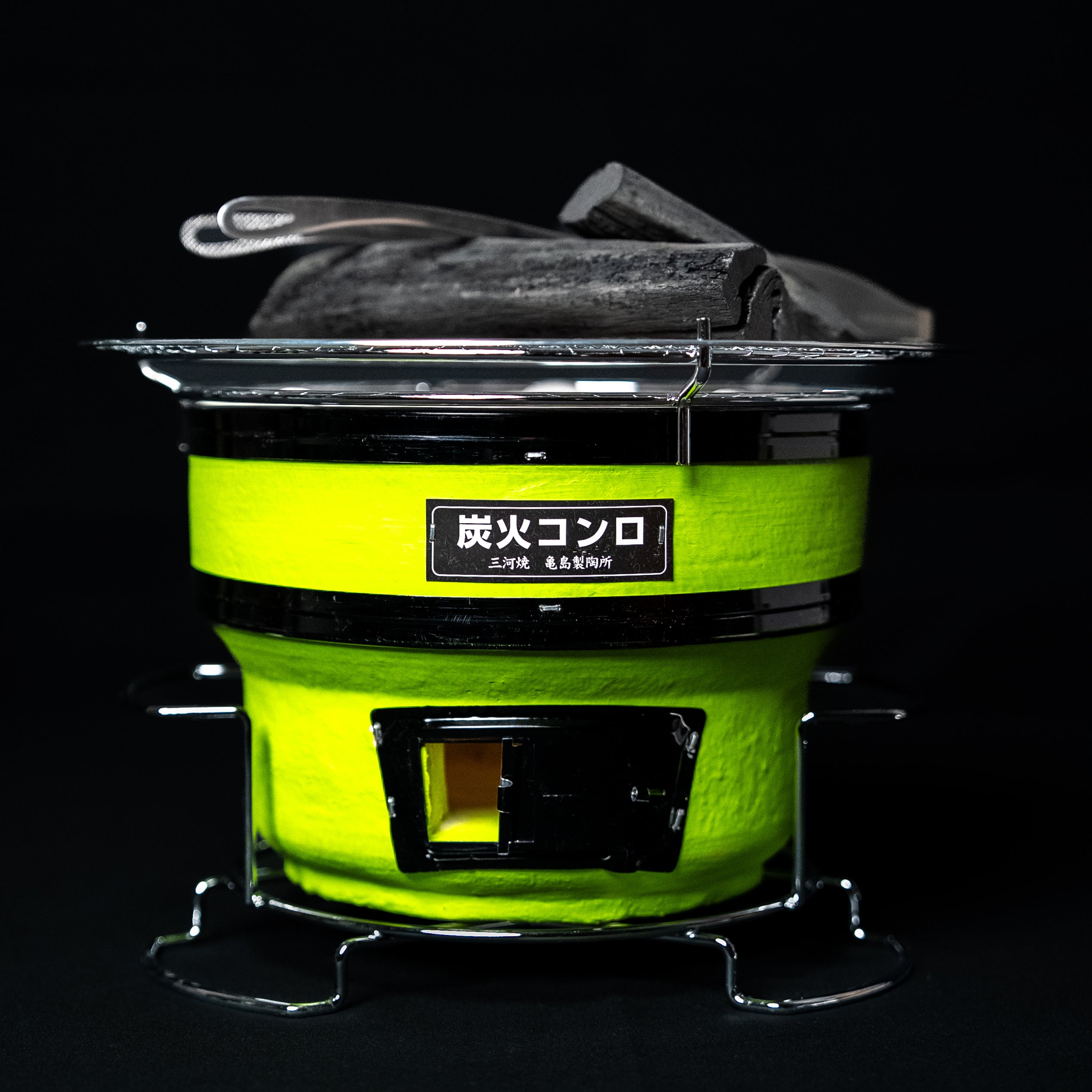 Small round Japanese charcoal grill in bright green with black accents, metal grate, and charcoal pieces on top, against a dark background.