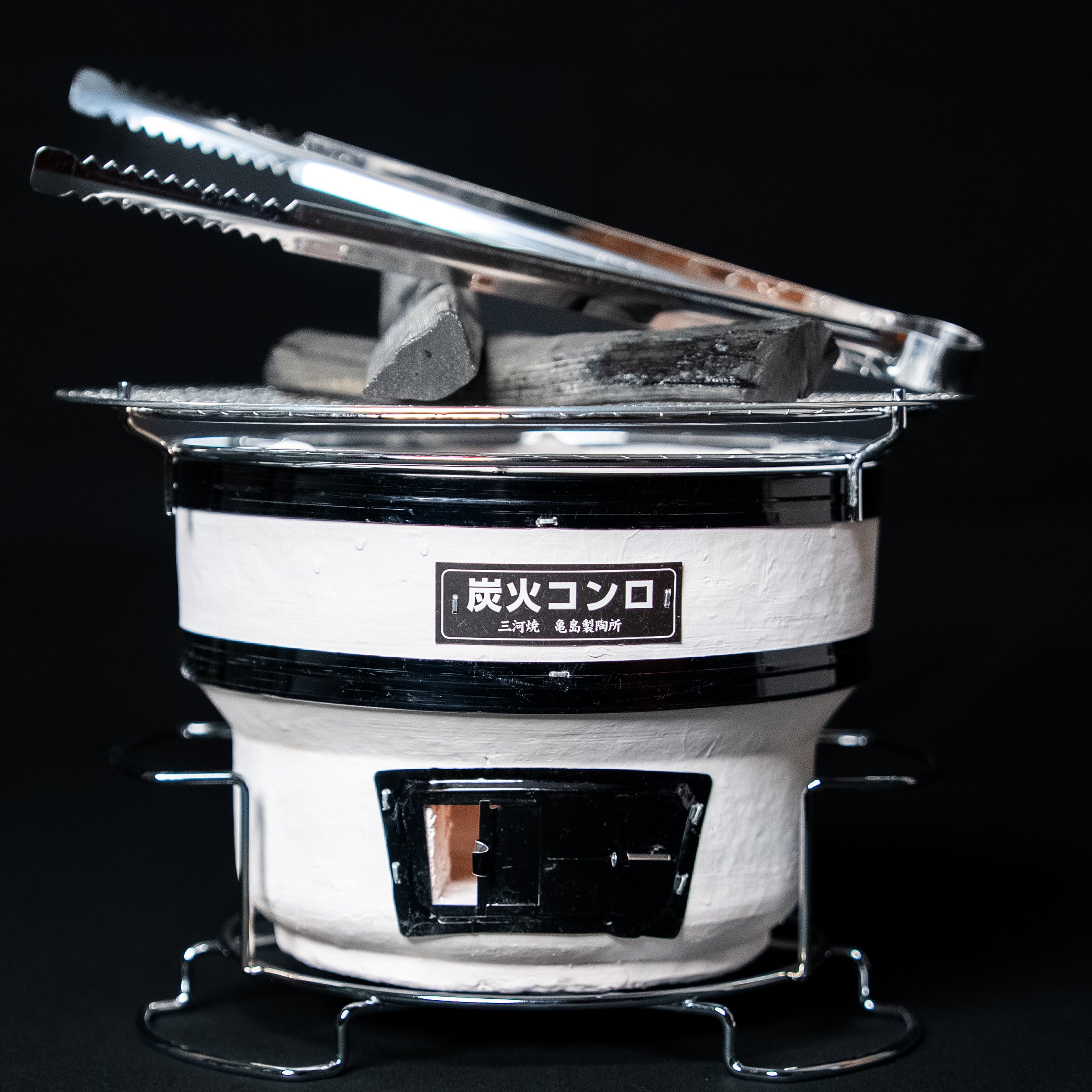 Small round Japanese charcoal grill in white with black accents, topped with charcoal and metal tongs, shown against a dark background.