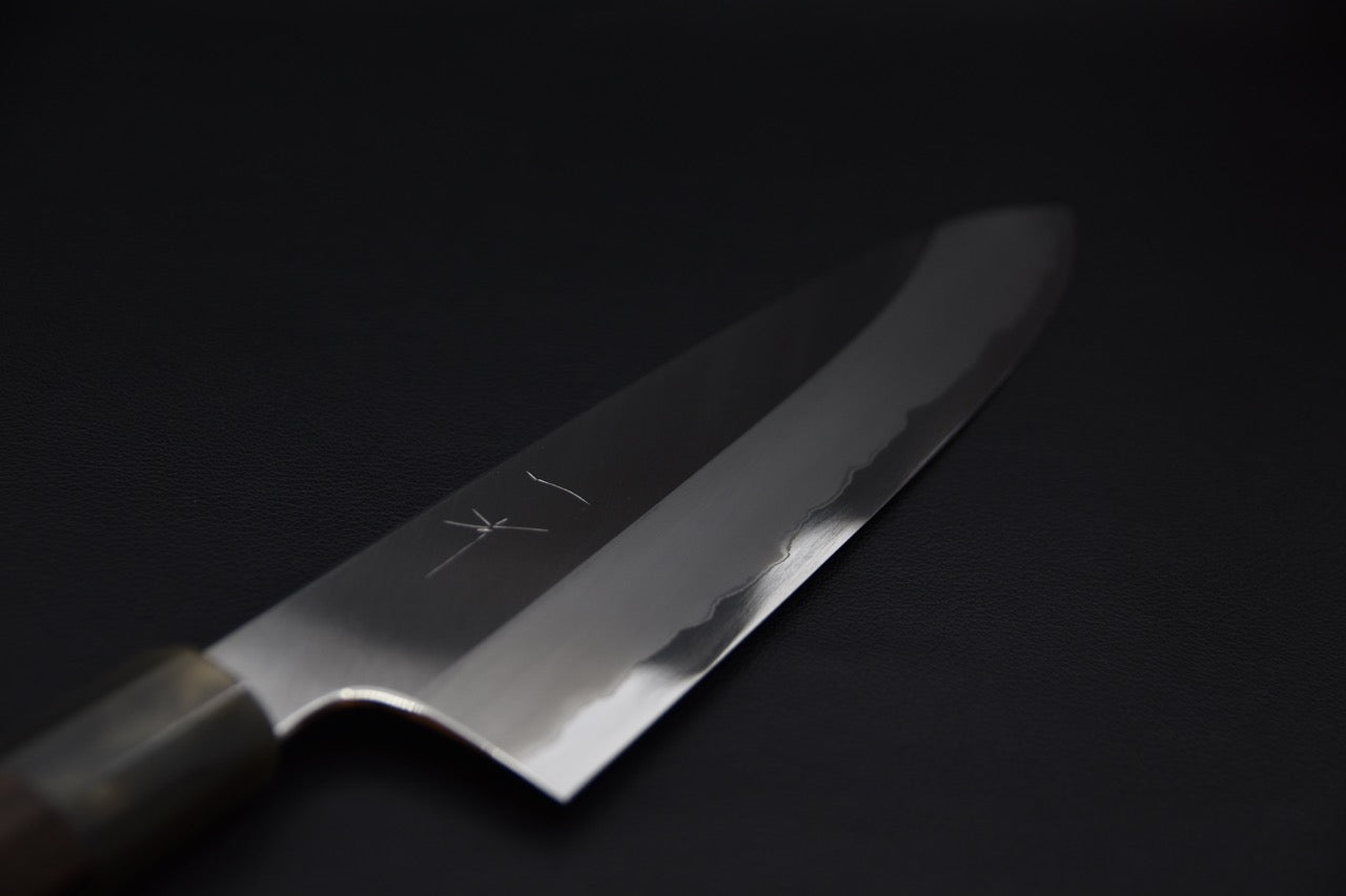 Close-up of a Japanese chef knife blade with polished finish and visible hamon line, shown against a dark background.