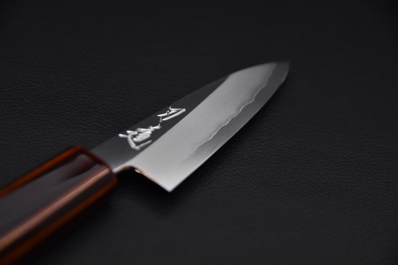 Close-up of a Japanese paring knife showing its polished blade and wavy hamon pattern, with a lacquered wooden handle, on a black background.