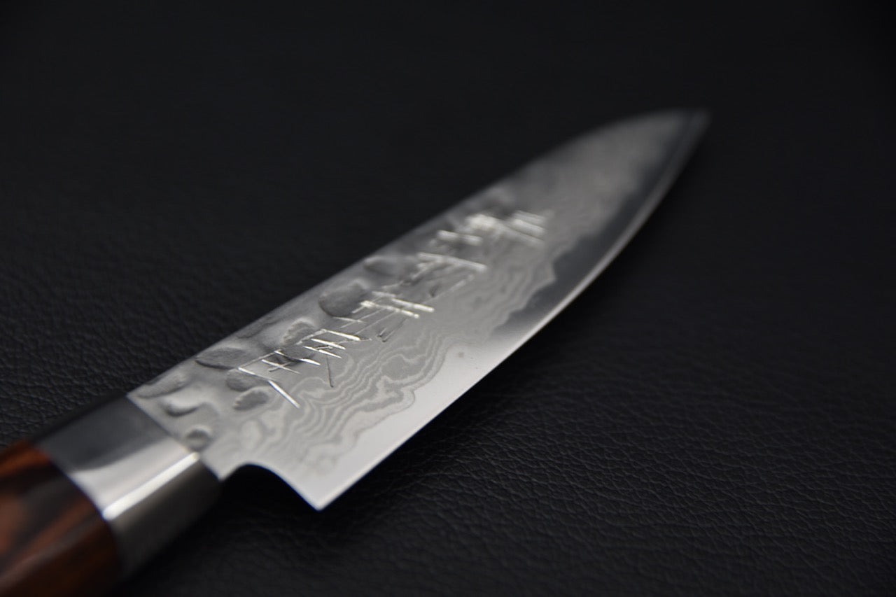 Close-up of a Japanese paring knife blade featuring a tsuchime hammered finish and Damascus pattern, shown against a black background.