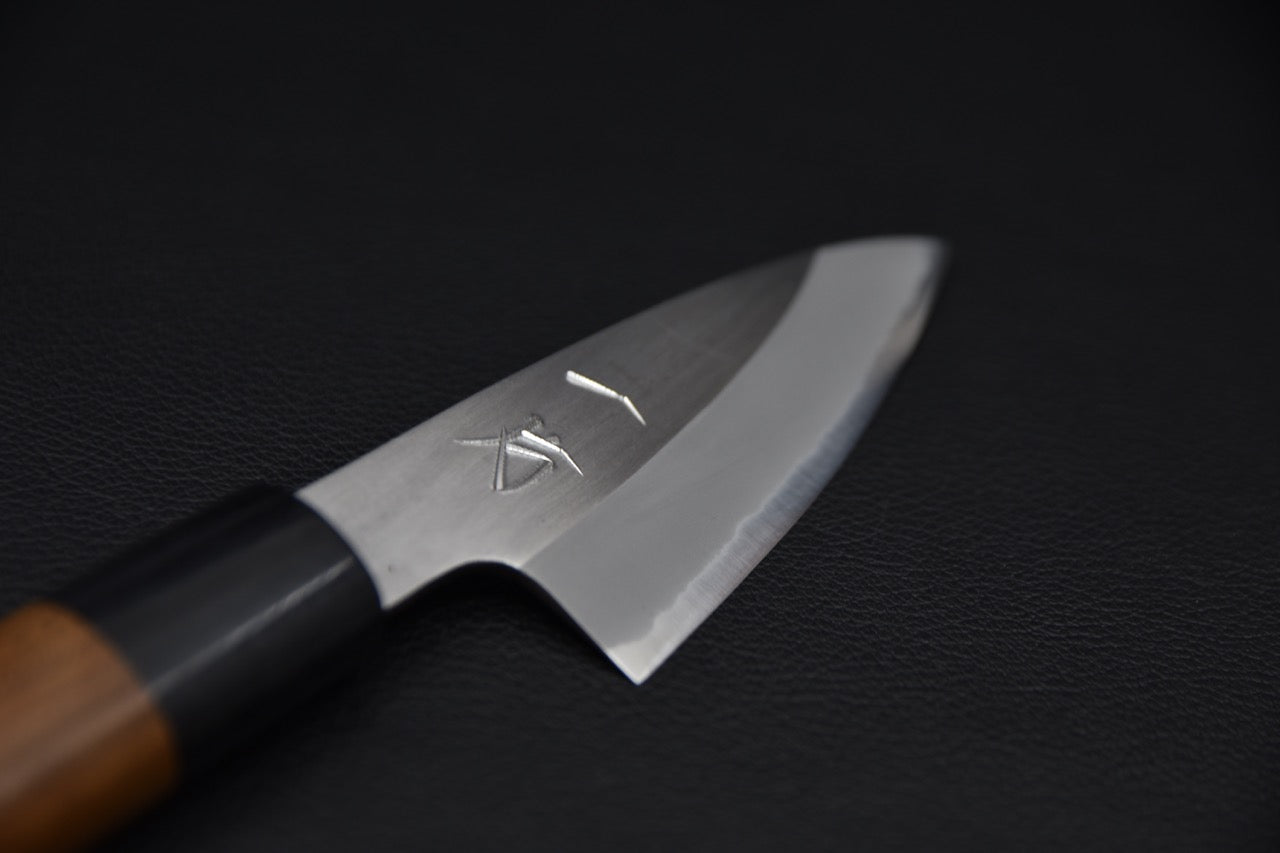 Close-up of a Japanese paring knife blade with engraved markings, featuring a polished edge and wooden handle, displayed on a black background.