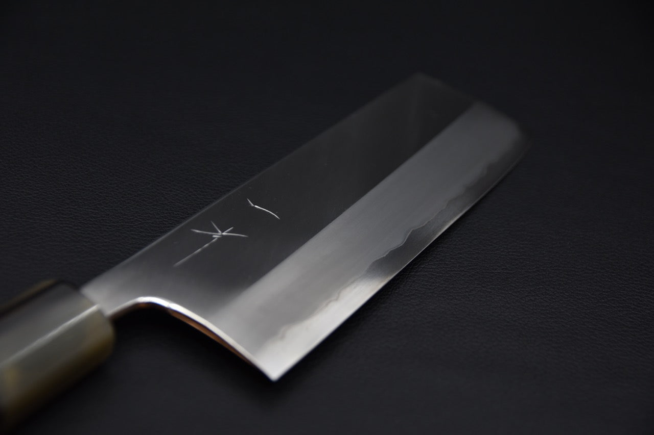 Close-up of a Japanese nakiri knife blade with a polished finish and visible hamon line, shown against a dark background.