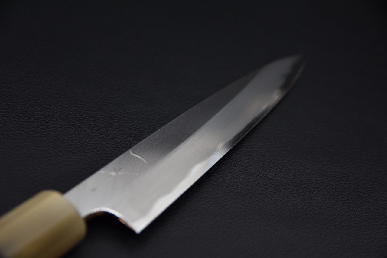 Close-up of a Japanese petty knife blade with a polished finish and subtle hamon line, shown against a dark background.