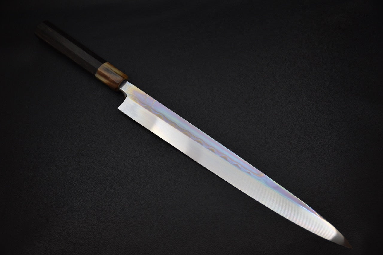 Japanese yanagiba knife with a long, single-edge polished blade and a dark wood (Macassar ebony) wa-style handle, showcased on a plain dark background.