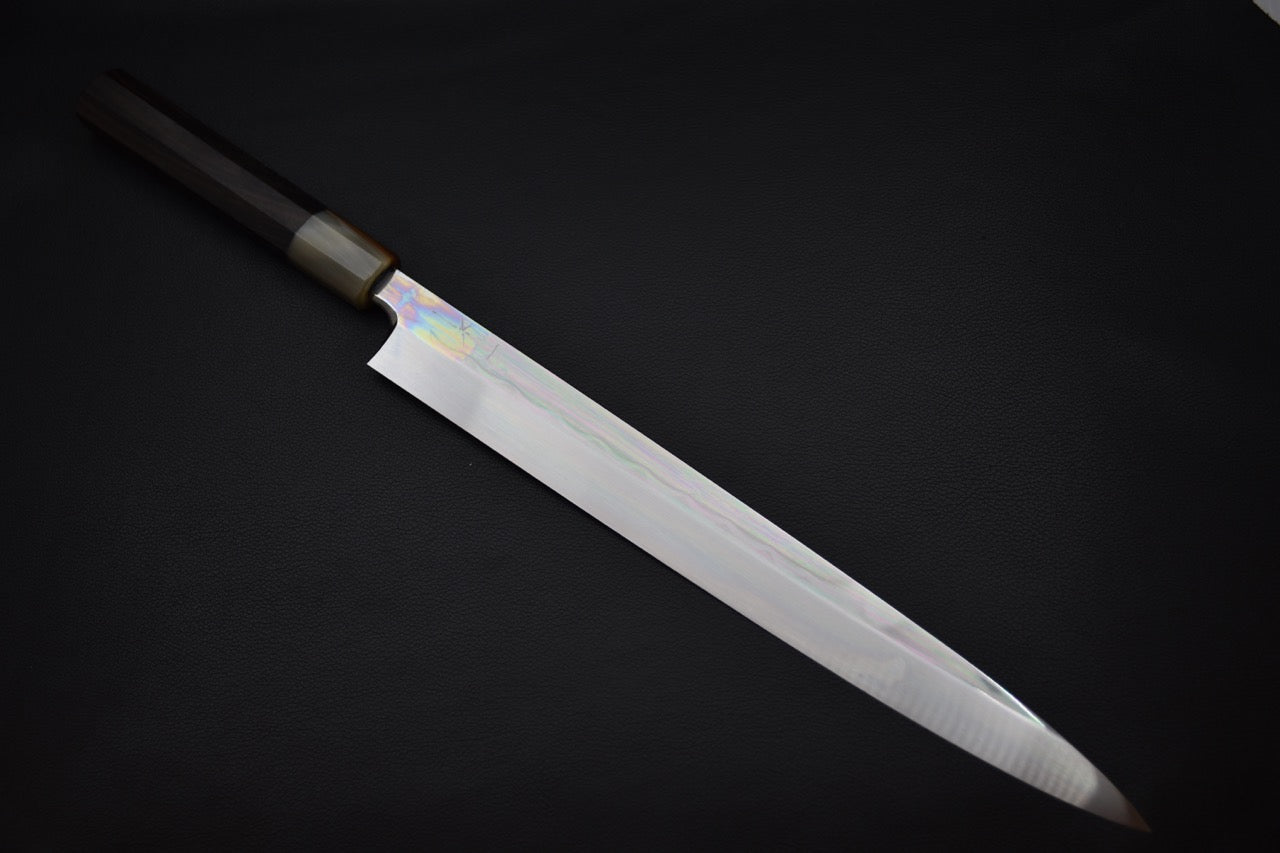 Japanese yanagiba knife with a long polished blade and a light-colored wa-style handle, displayed on a dark background.