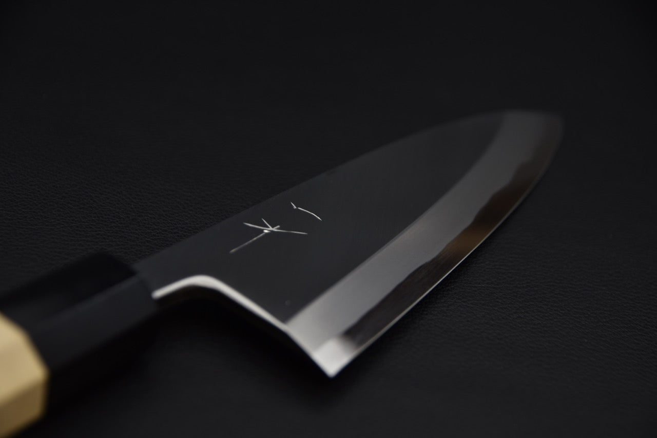 Close-up of a Japanese deba knife blade with a polished finish and engraved kanji, shown against a dark background.