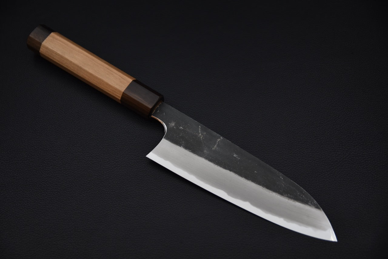 Japanese santoku knife with a kurouchi finish and polished cutting edge, featuring a wooden wa-handle, displayed on a dark background.
