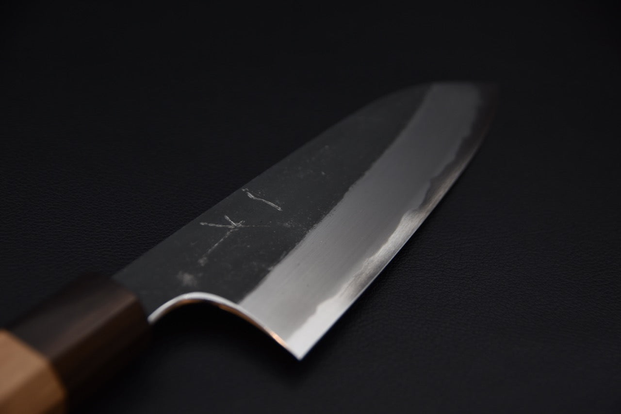 Close-up of a Japanese santoku knife blade with a kurouchi finish and polished cutting edge, displayed on a dark background.