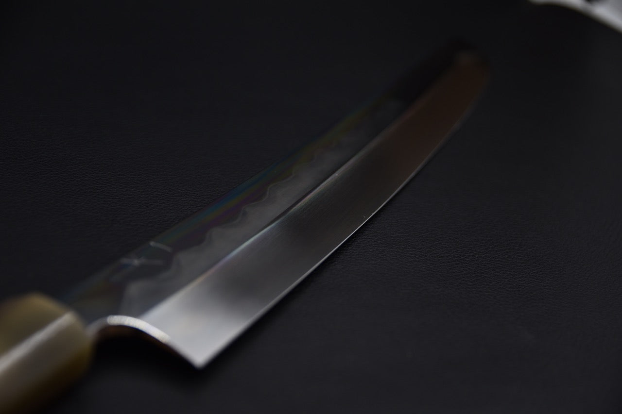 Close-up of a Japanese sakimaru yanagiba knife showing a long polished blade with a subtle hamon line and a dark wa-style handle, set against a dark background.