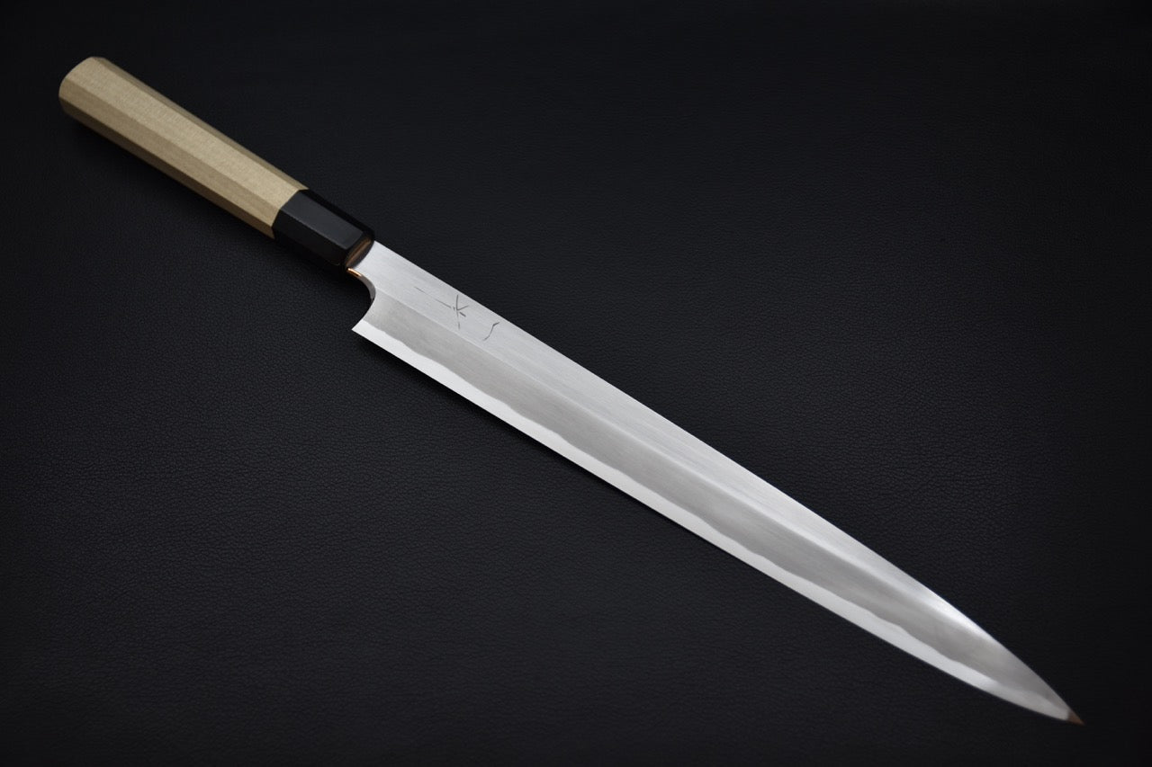 Japanese yanagiba knife with a long polished blade and light wooden wa-handle, displayed on a dark background.