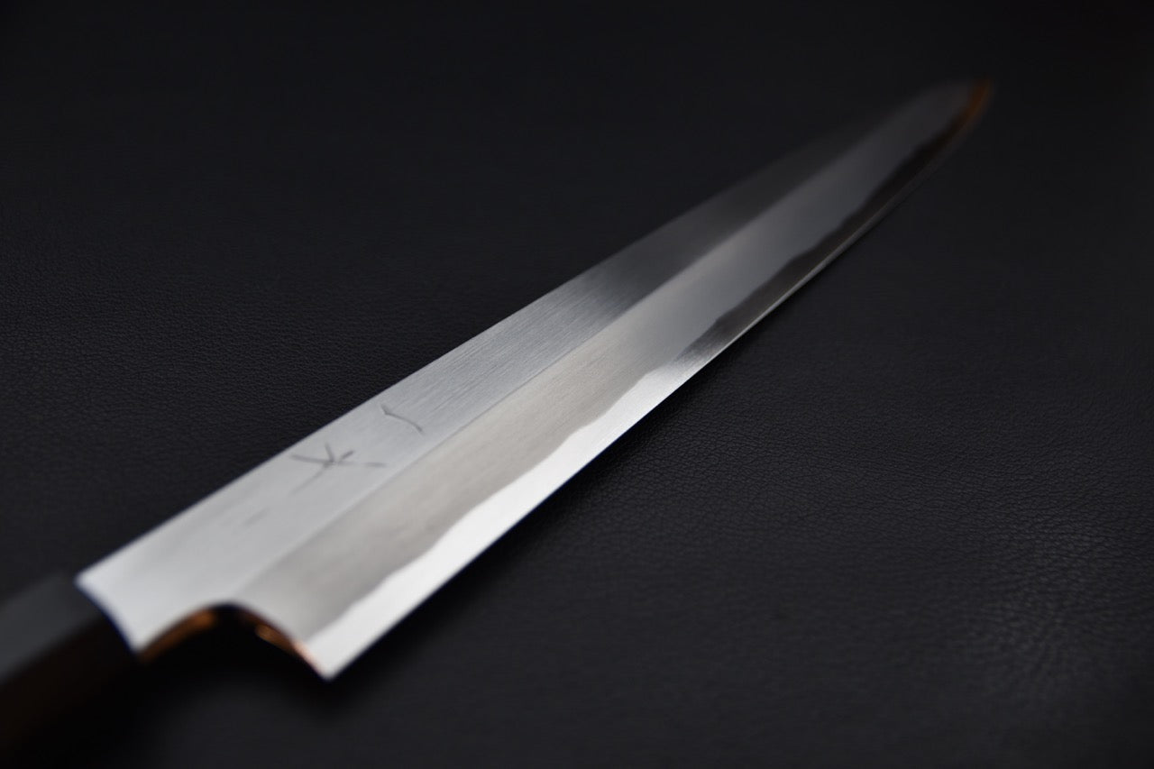 Close-up of a Japanese yanagiba knife blade with a polished finish and visible hamon line, displayed against a dark background.
