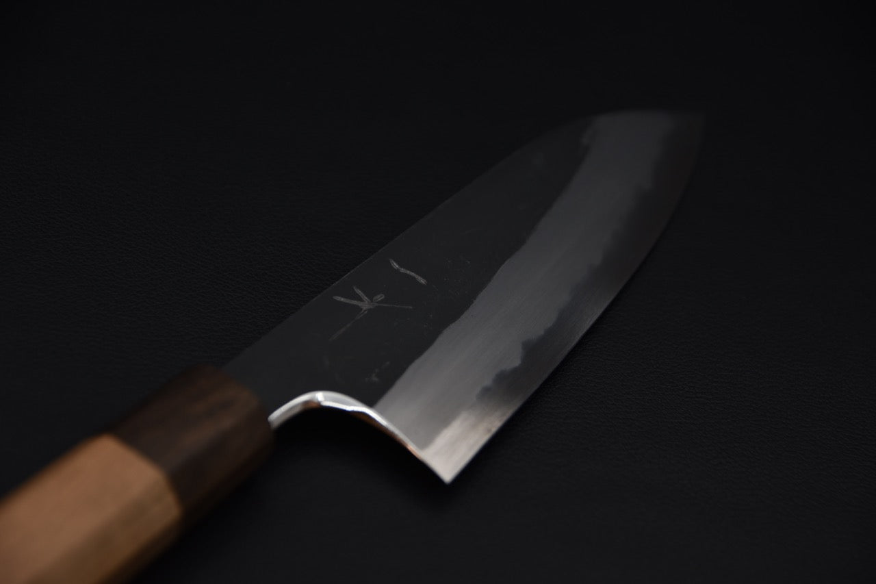 Close-up of a Japanese santoku knife blade with a polished cutting edge and kurouchi finish, displayed against a dark background.