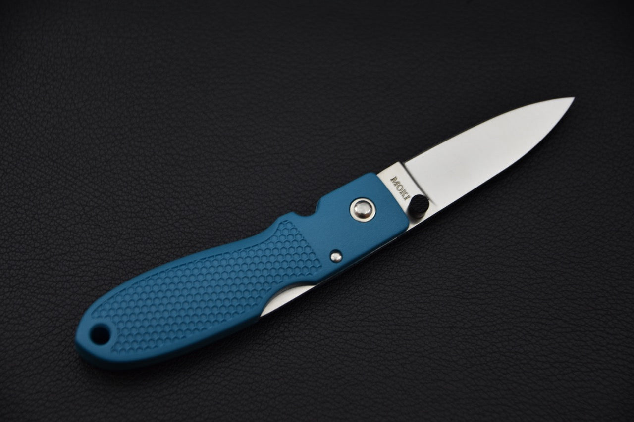 Inscription "MOKI" visible près du talon de la lame, couteau ouvert sur fond noir.