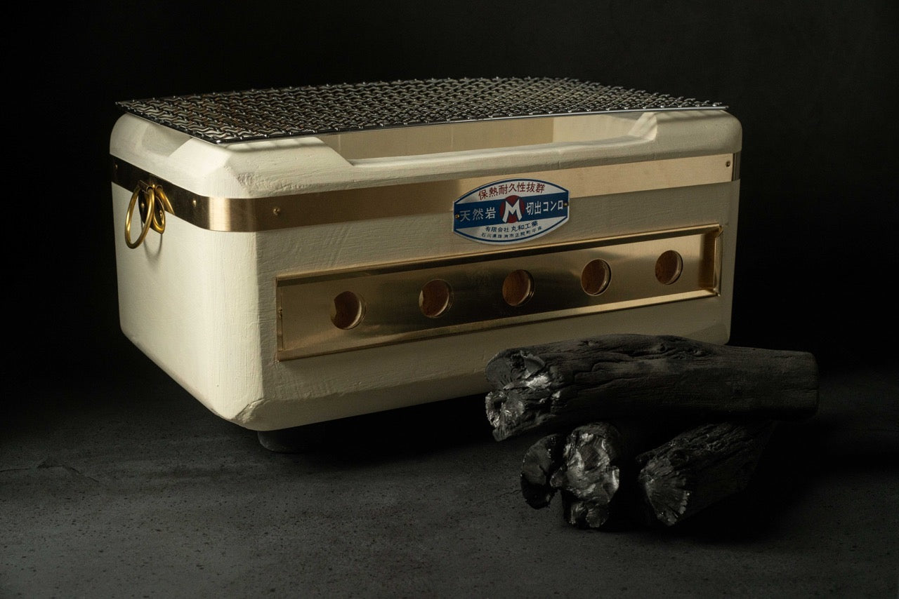 Japanese rectangular charcoal grill in cream color with gold accents, mesh top, and side handles, shown with charcoal pieces on a dark background.