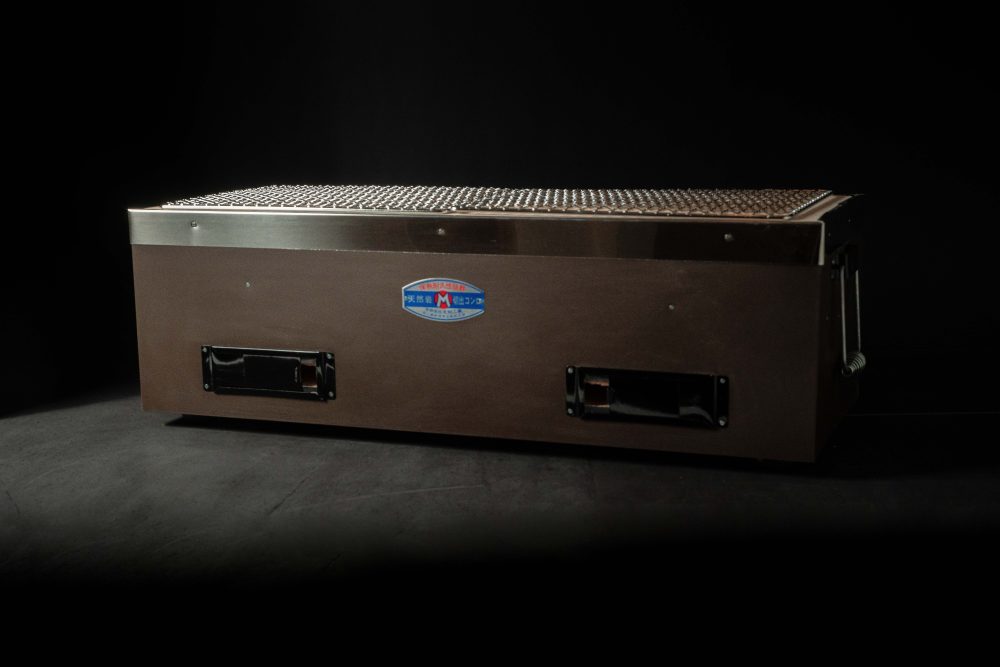 Large rectangular Yakitori Konro charcoal grill with mesh grate and blue Japanese label, placed on a dark surface against a black background.
