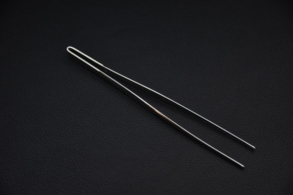 Long stainless steel cooking tweezers with a slim, looped handle design, displayed on a dark textured background.