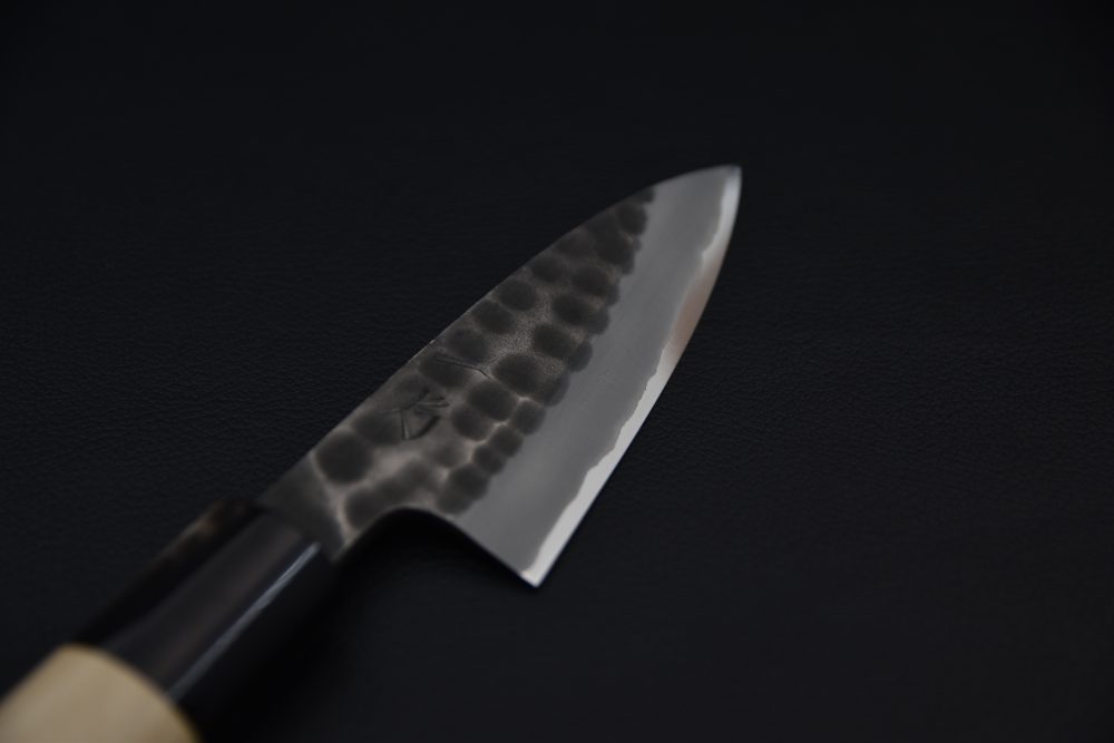 Close-up of a Japanese knife blade featuring a hammered tsuchime finish and polished edge, shown against a dark background.