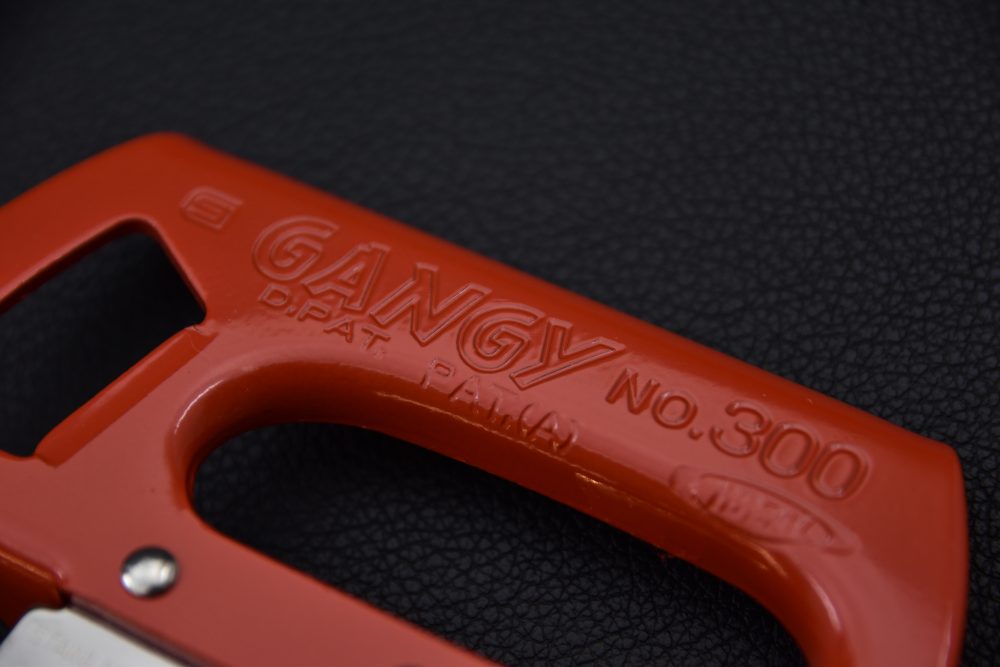 Close-up of an orange plastic handle with ‘GANGY NO.300’ embossed on it, on a dark background.