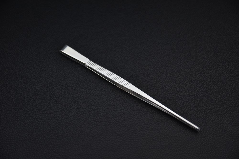 Slim stainless steel tweezers featuring a narrow tip and ridged grip section, displayed on a dark textured background.