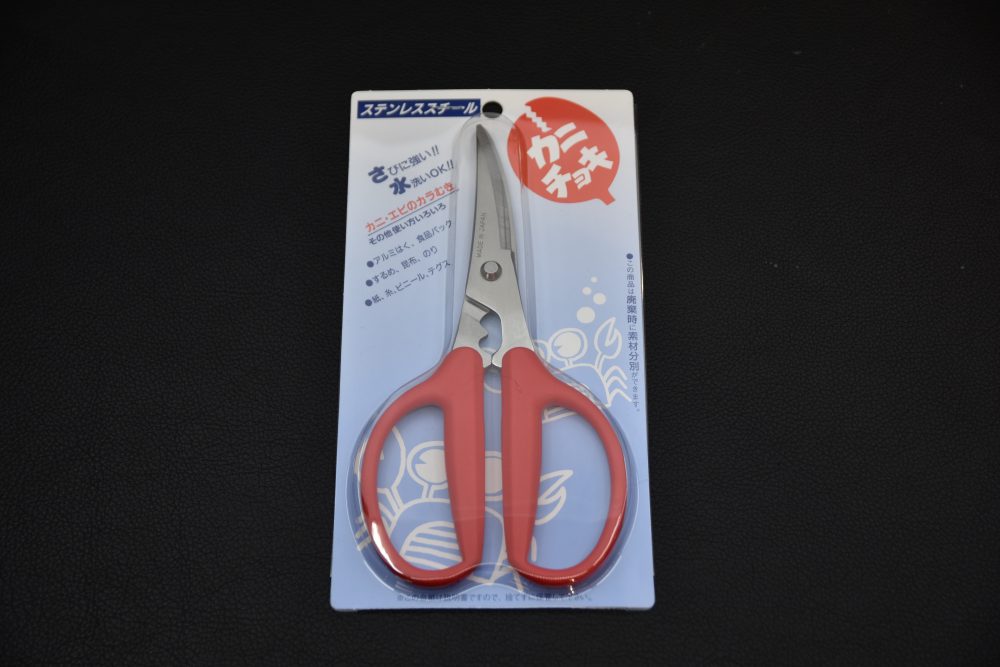 Japanese stainless-steel kitchen scissors with red handles, shown in their original packaging on a dark background.
