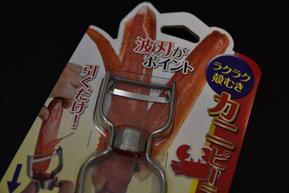 Japanese crab leg peeler in its retail packaging, featuring metal blade and illustrated instructions, shown against a dark background.