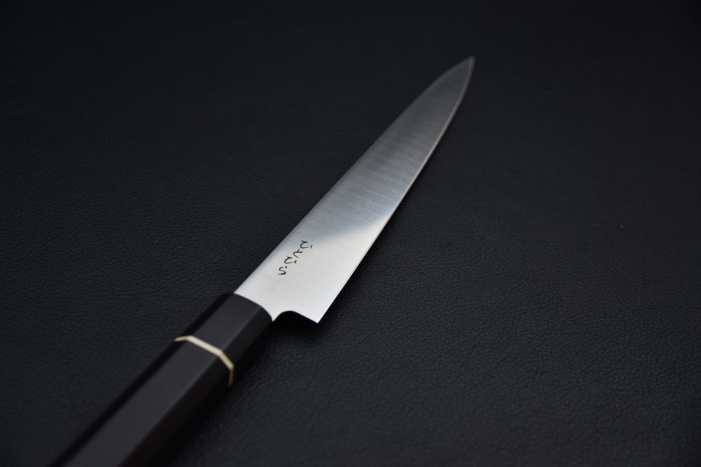 Japanese utility knife with a sharp stainless steel blade, dark wooden handle, and metal ferrule on a black textured background.