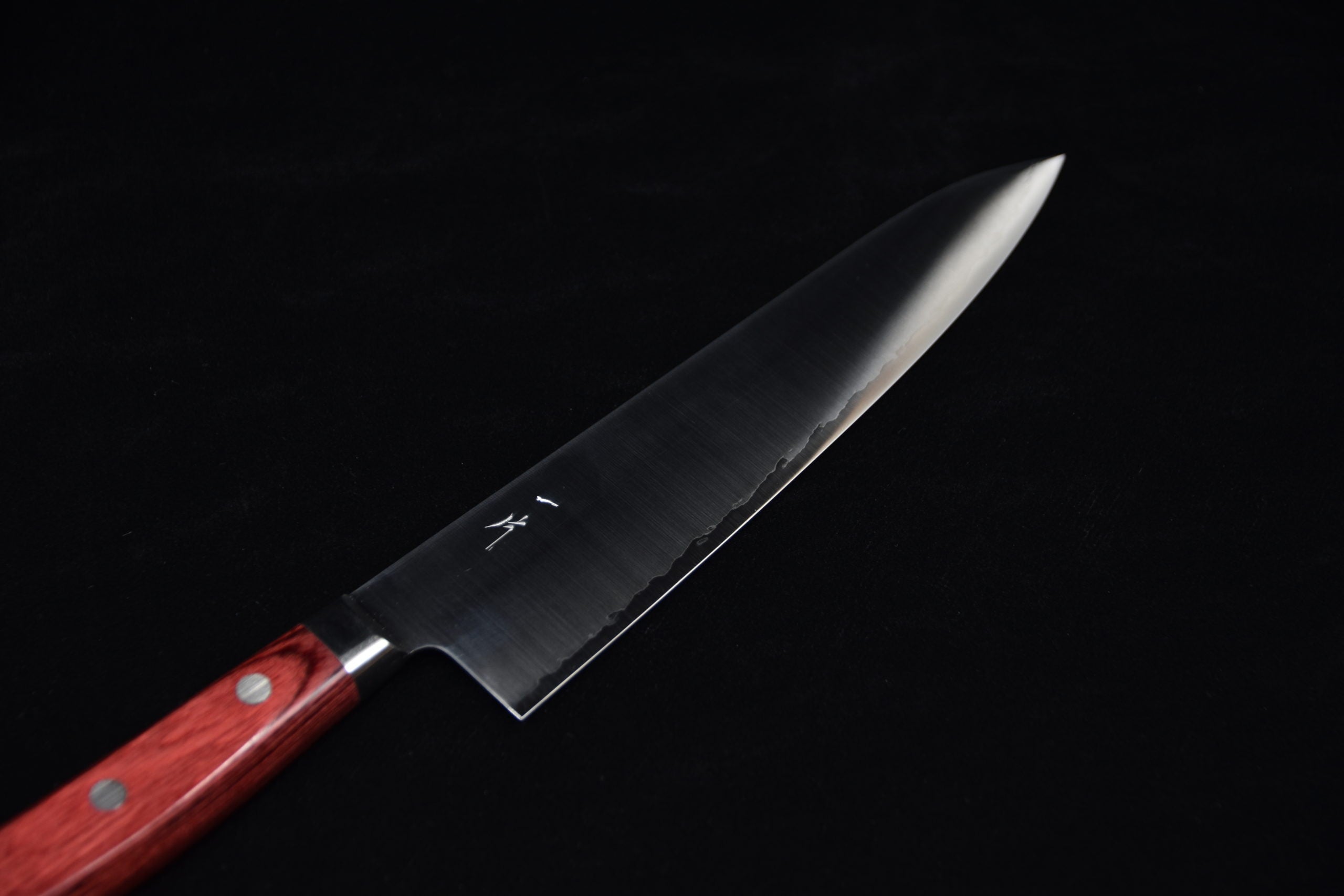 Japanese chef knife with a long stainless steel blade and a red wooden handle with rivets, displayed on a black background.