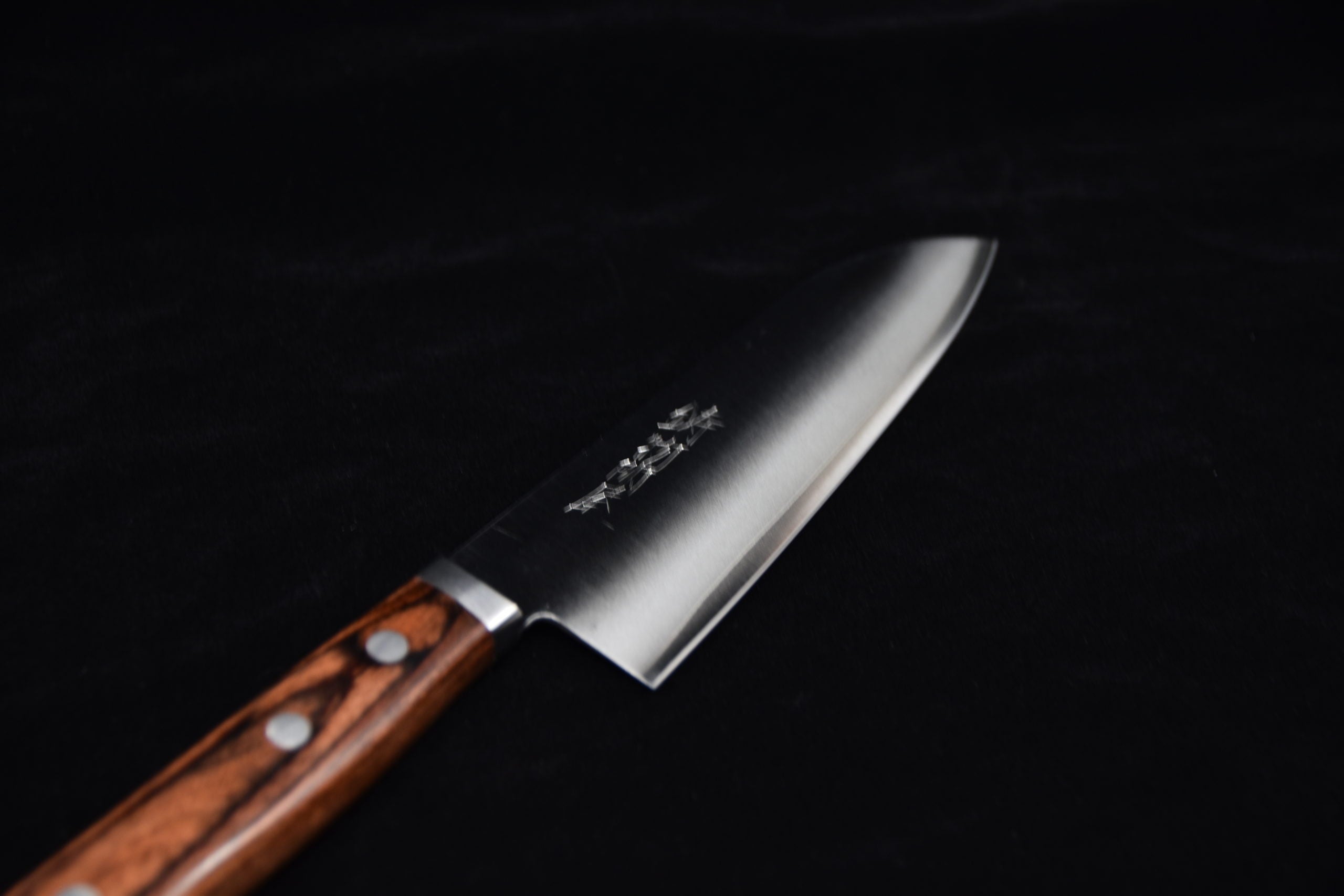 Japanese santoku knife with a wide stainless steel blade and a brown wooden handle with rivets, placed on a black background.