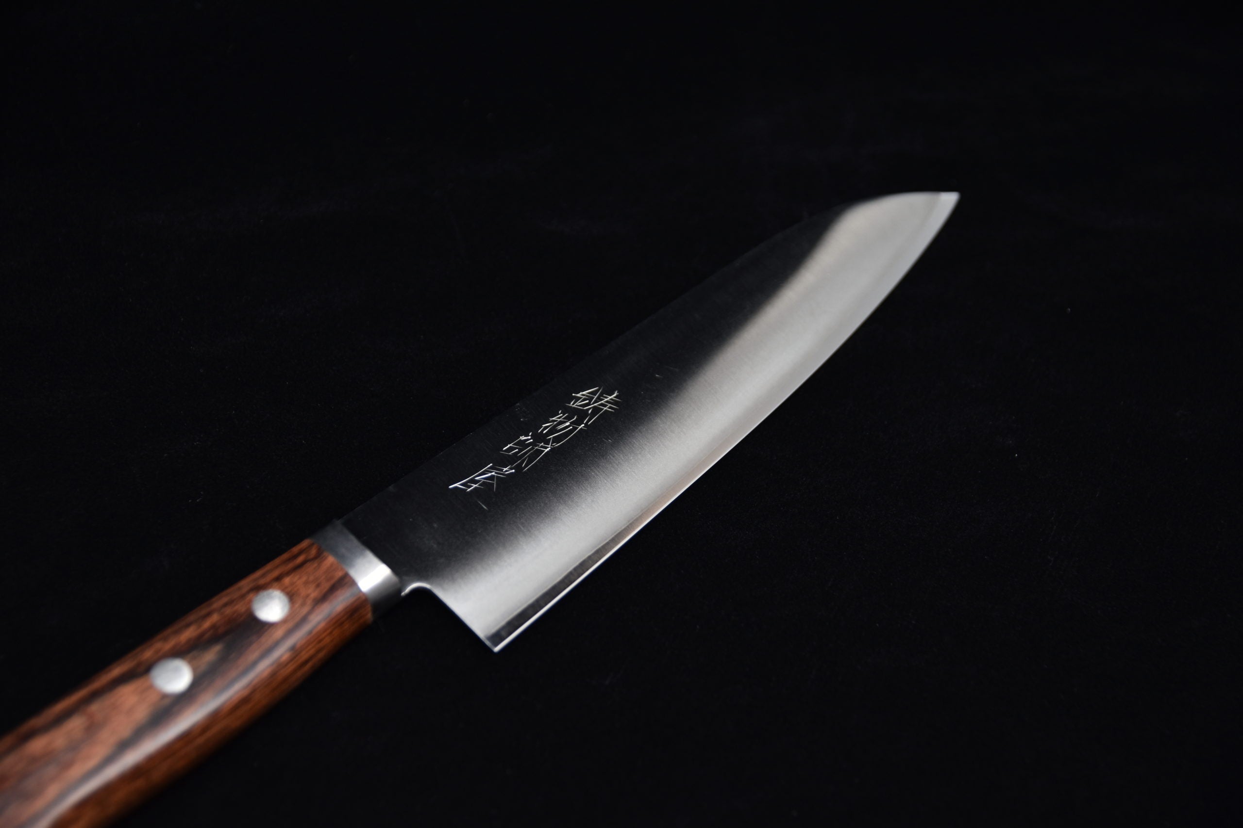 Japanese chef’s knife with polished steel blade and brown wooden handle, featuring engraved kanji characters, on a black background.