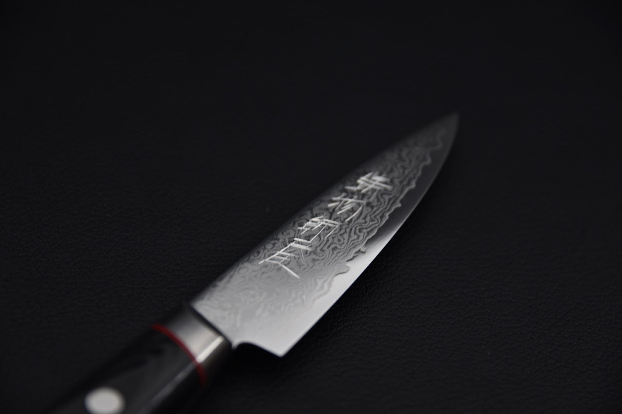 Close-up of a Japanese paring knife blade with a detailed Damascus pattern and engraved kanji, shown against a dark background.