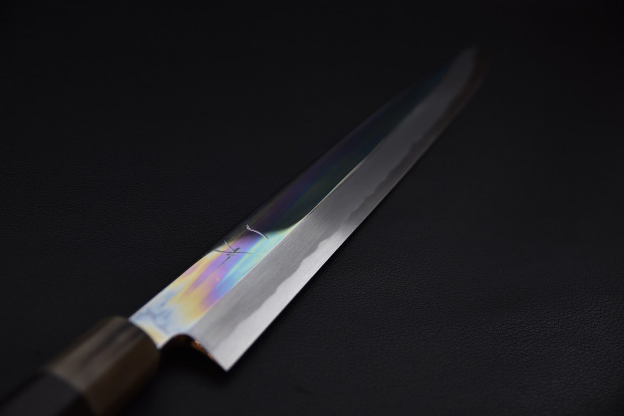 Close-up of a Japanese yanagiba knife blade with polished finish and subtle hamon line, shown against a dark background with a dark wooden wa-handle visible in the foreground.