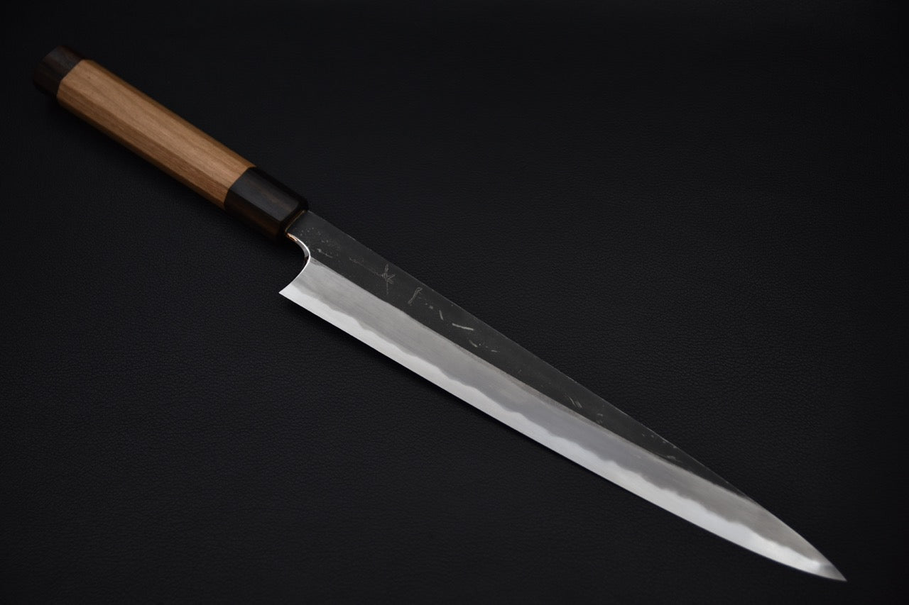 Japanese sujihiki knife with a long polished edge and kurouchi-finished spine, featuring a wooden wa-handle, displayed on a dark background.