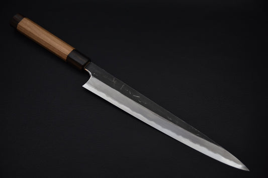Japanese sujihiki knife with a long polished edge and kurouchi-finished spine, featuring a wooden wa-handle, displayed on a dark background.