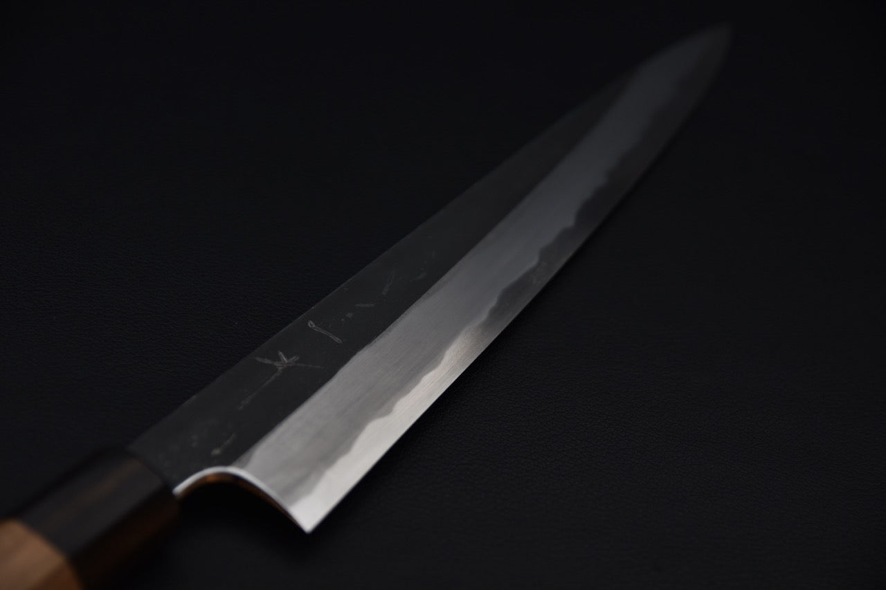 Close-up of a Japanese sujihiki knife blade with a polished cutting edge and kurouchi-finished spine, shown against a dark background.