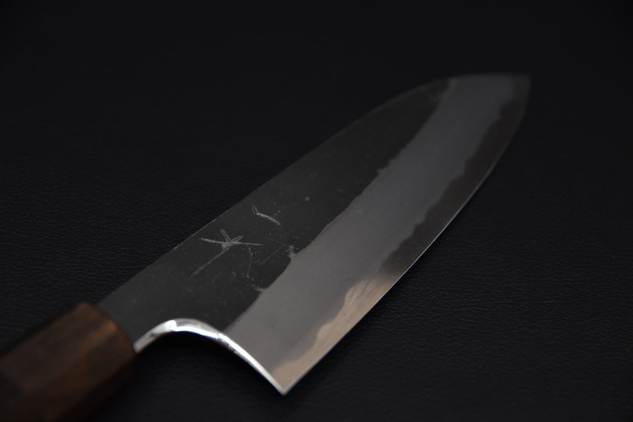 Close-up of a Japanese santoku knife blade with a kurouchi finish and polished edge, displayed on a dark background.