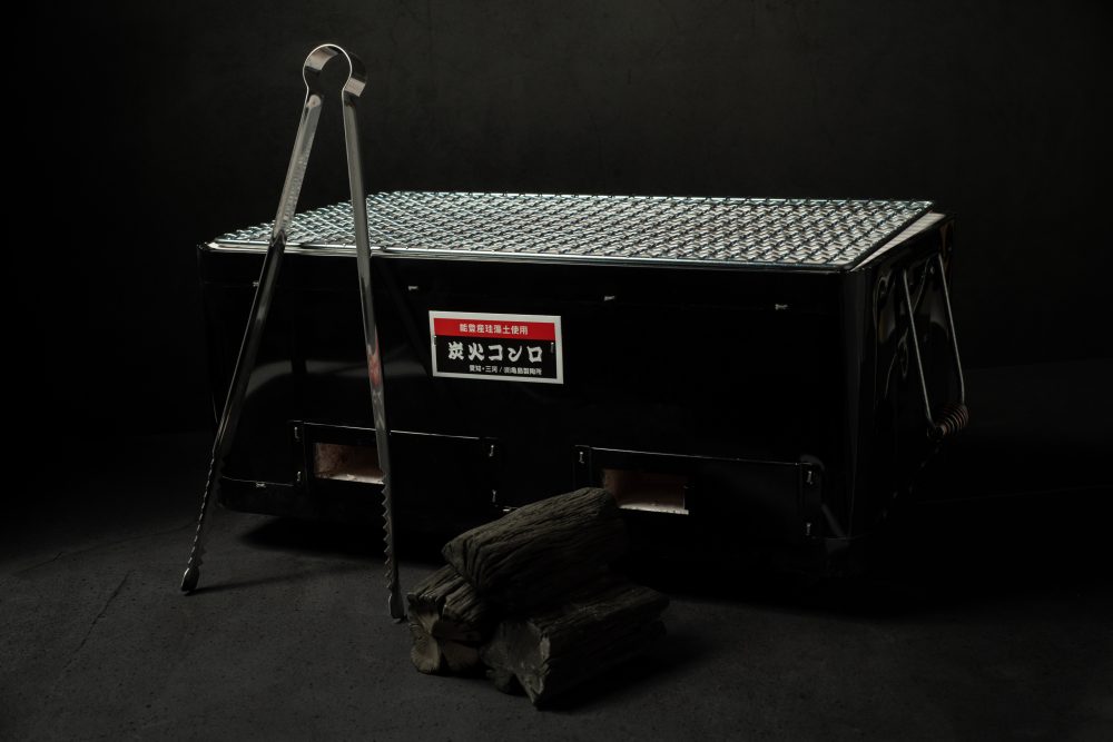 Black Yakitori Konro charcoal grill with mesh grate, long metal tongs, and stacked charcoal pieces on a dark surface and background.