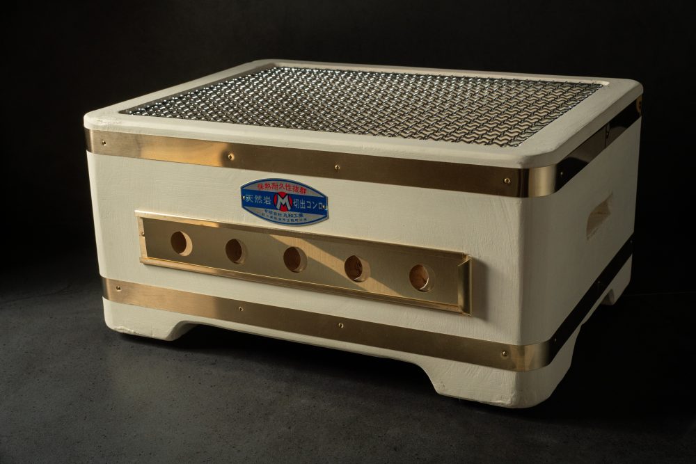 Yakitori Konro charcoal grill with brass trim, circular vents, and mesh grate, displayed on a dark surface against a black background.