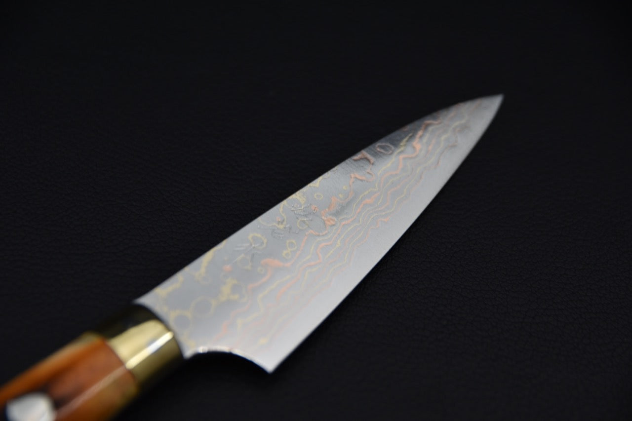Close-up of a Japanese petty knife blade featuring a rainbow Damascus pattern with layered waves, displayed against a dark background.
